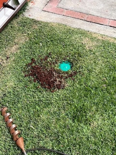 Sentricon® bait station installation in a grassy yard area, with surrounding soil and drill auger visible.
